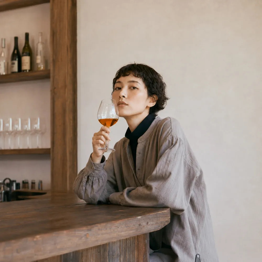 ち着いたトーンのバーカウンターに肘をつき、美しく輝くビネヒ（Binekhi）のオレンジワインが入ったグラスを手に、こちらを見つめるショートヘアの女性モデル
