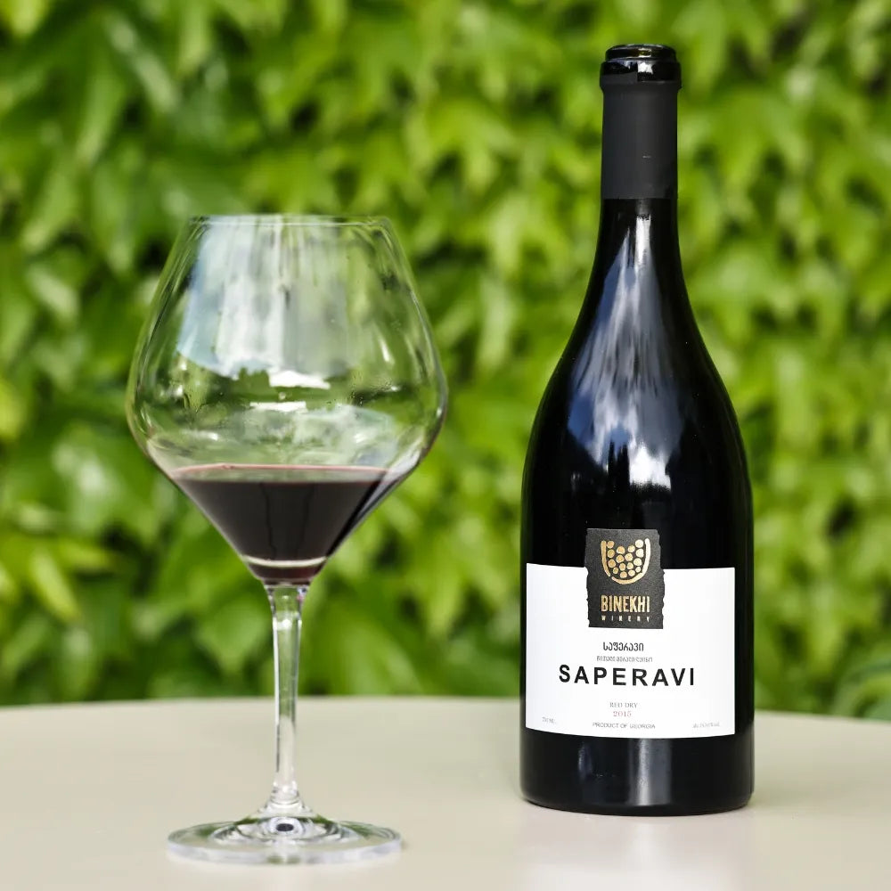 鮮やかなグリーンの葉をバックに、白いテーブルの上に美しく配置された「BINEKHI SAPERAVI（ビネヒ サペラビィ）」のワインボトルと赤ワイン入りのグラス