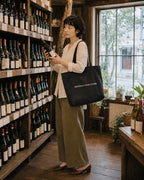 ギフトのワインのラベルを確かめる女性、ブラックのトートバッグで窓辺の店内